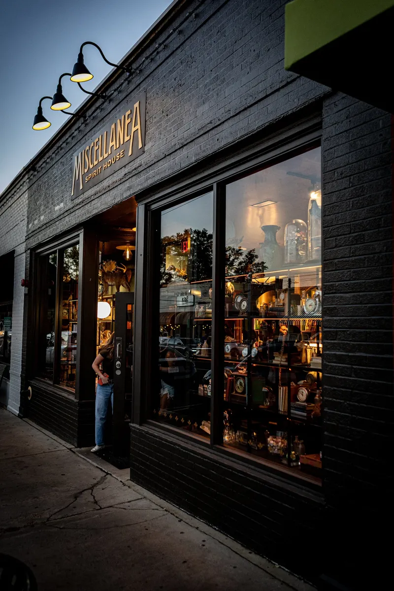 The black brick front of Miscellanea Spirit House at dusk, with its name on the facade and shelves of clocks and curios in the windows.